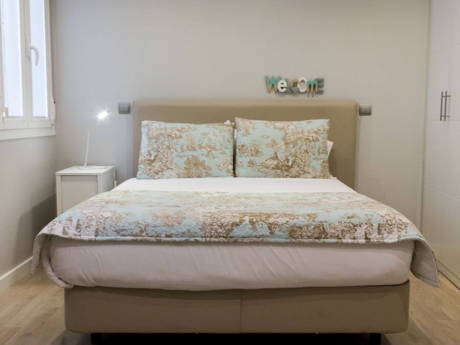 Un dormitorio con cama, mesita de noche, lámpara y un cartel que dice 'Welcome'.