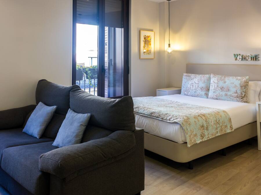 A modern bedroom with a sofa, a bed with floral bedding, a bedside table, and a wall-mounted light fixture.