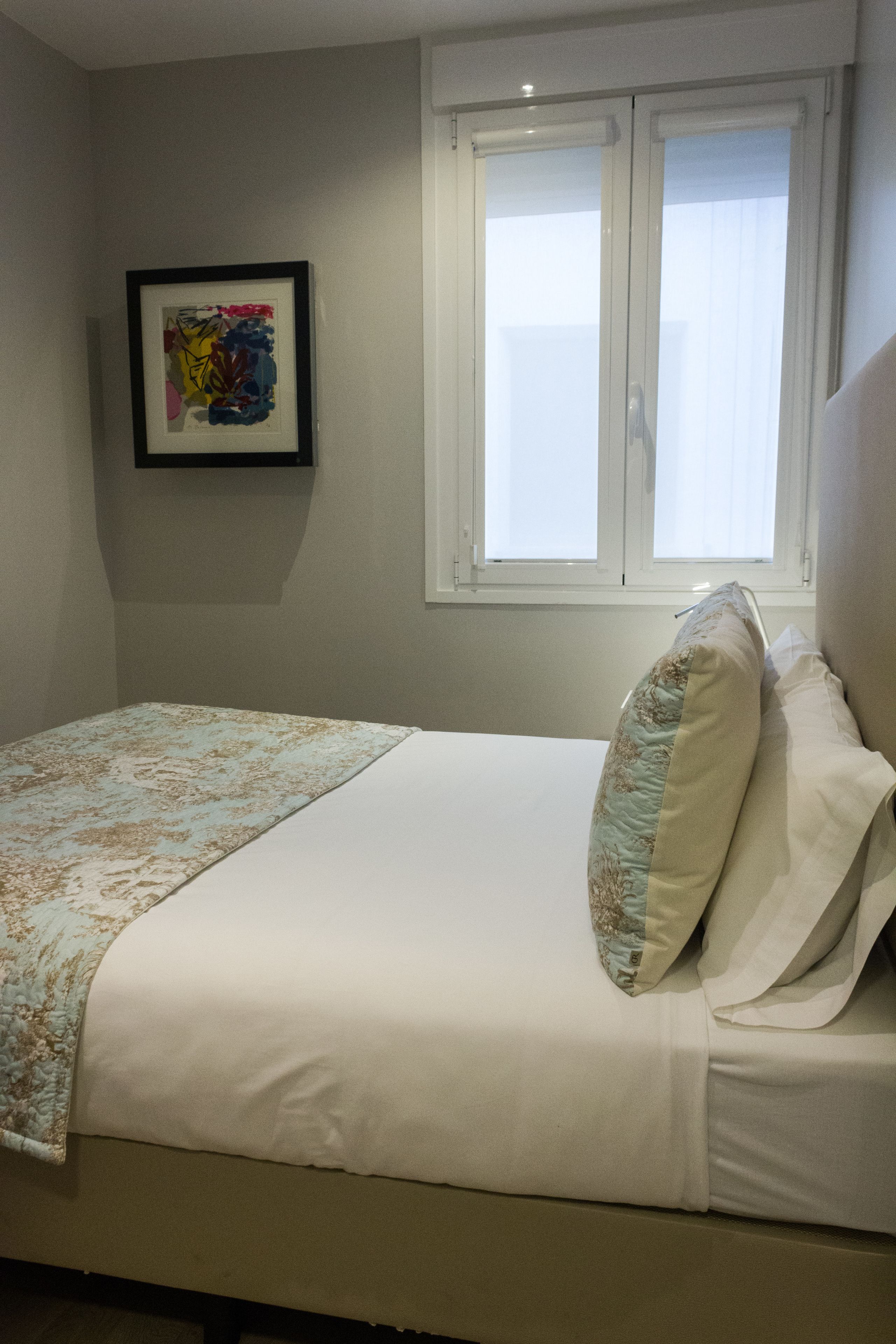 A neatly made bed with a patterned bedspread and pillows, a framed abstract artwork on the wall, and a window with curtains.