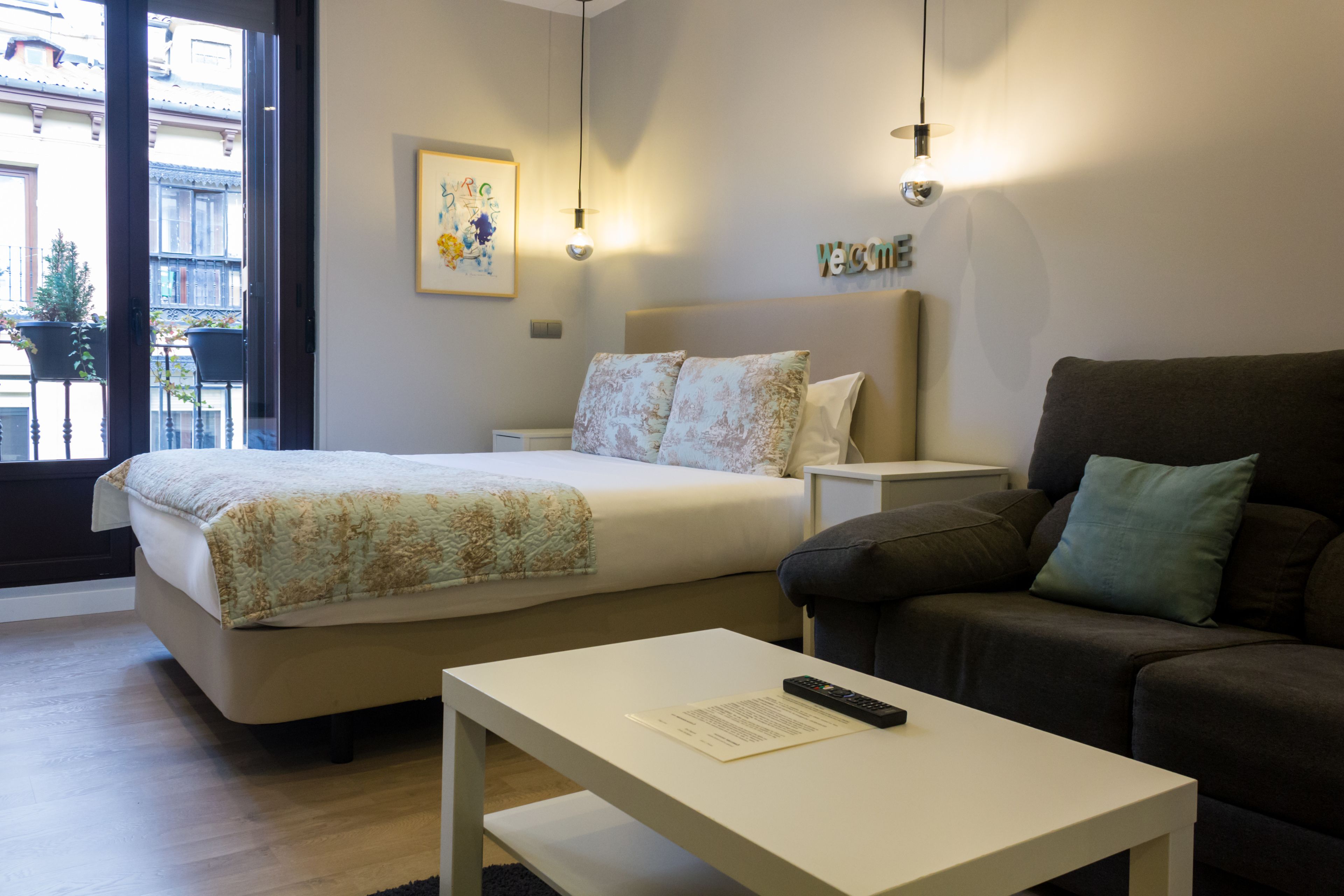 A hotel room with a bed, a bedside table, a couch, and a window with a view of buildings.