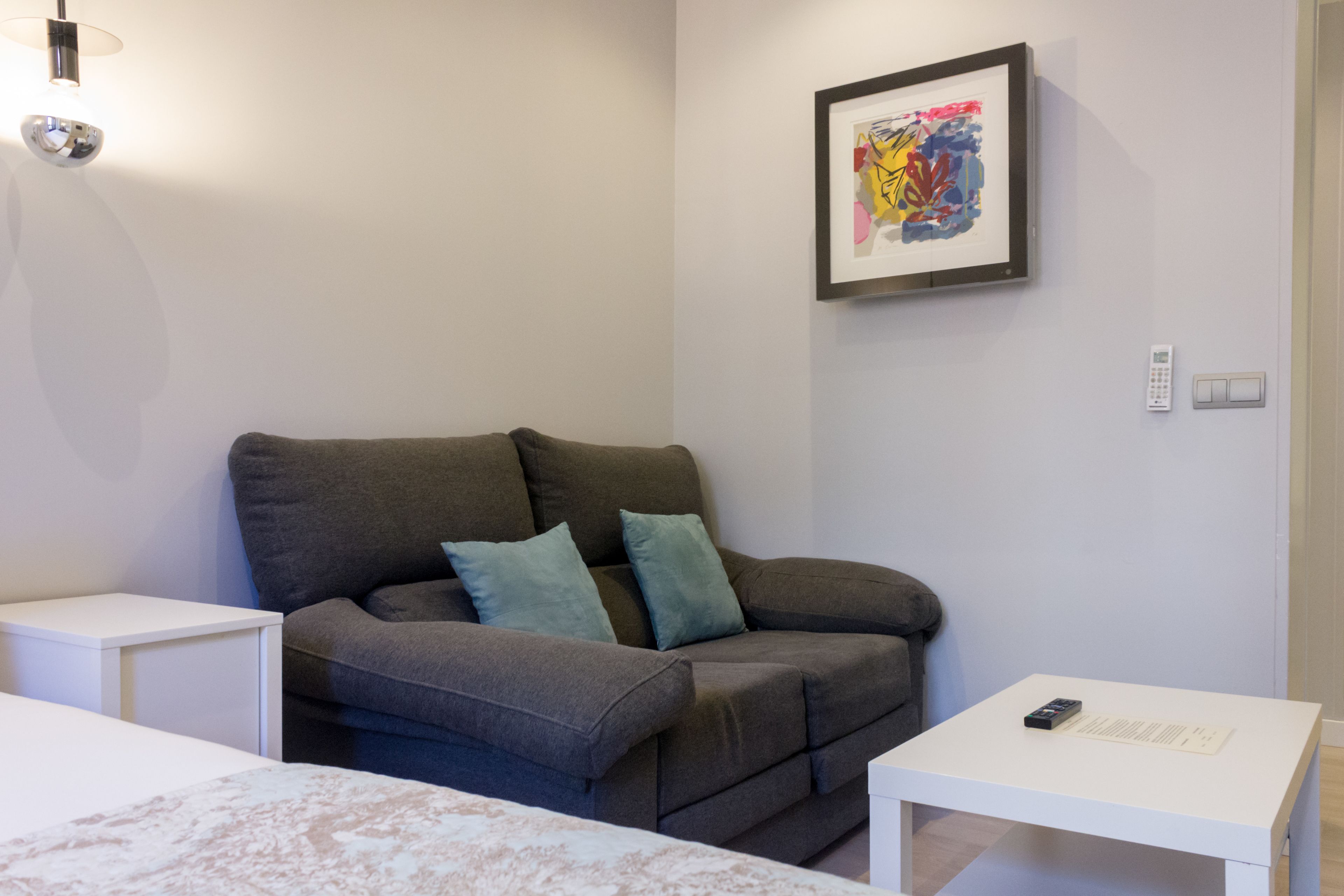 A modern living room with a grey sofa, two teal cushions, a white side table, a white coffee table, and a framed abstract painting on the wall.