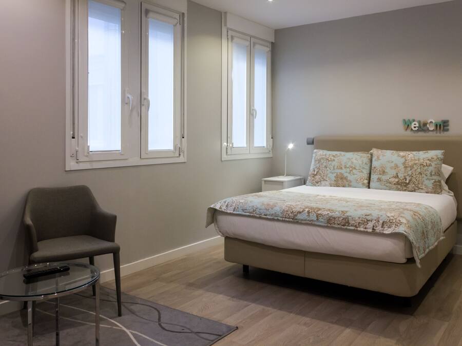 Un dormitorio con una cama, una silla, una mesita y dos ventanas.