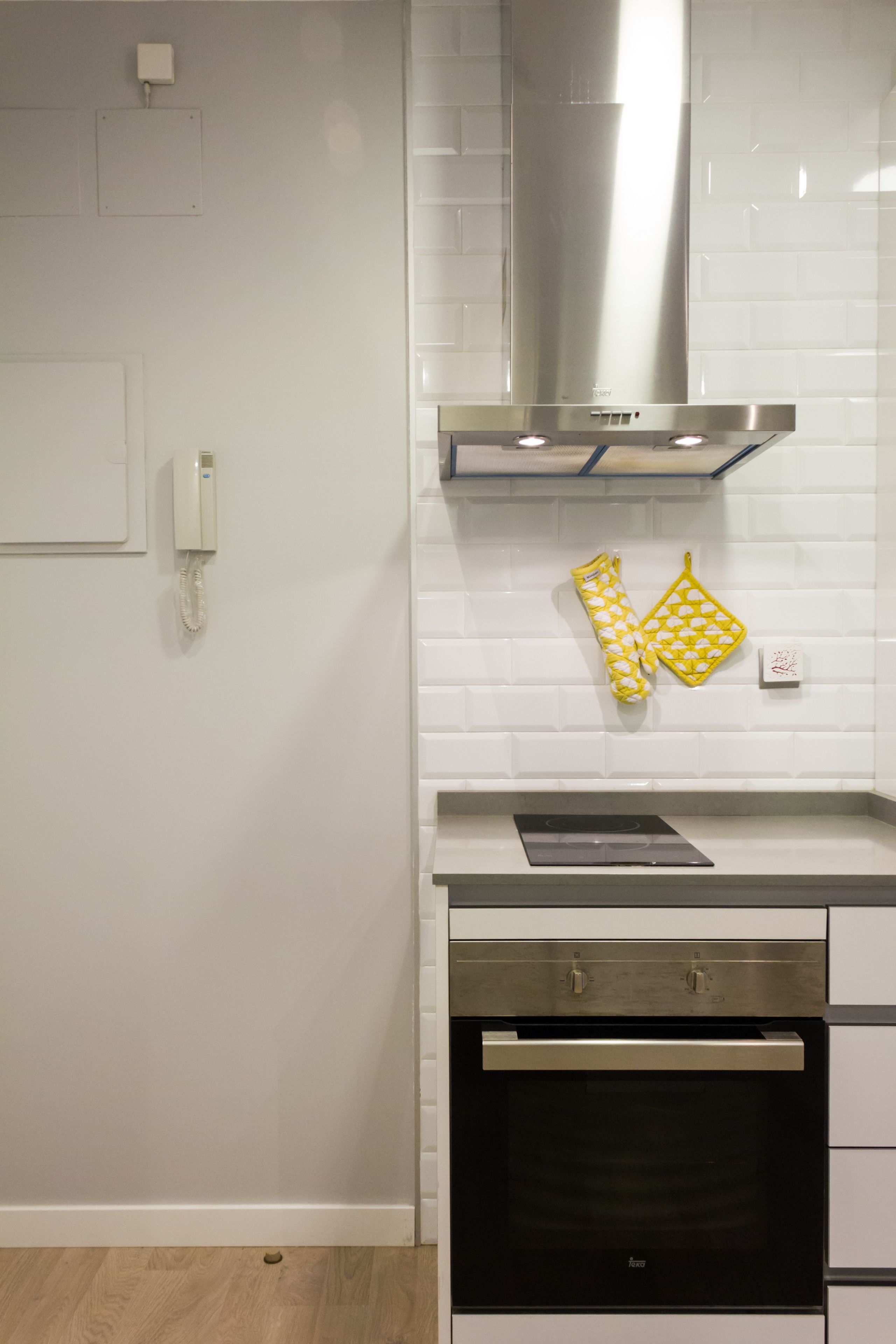 Una cocina con paredes de azulejos blancos, extractor de acero inoxidable, horno empotrado y un teléfono en la pared.