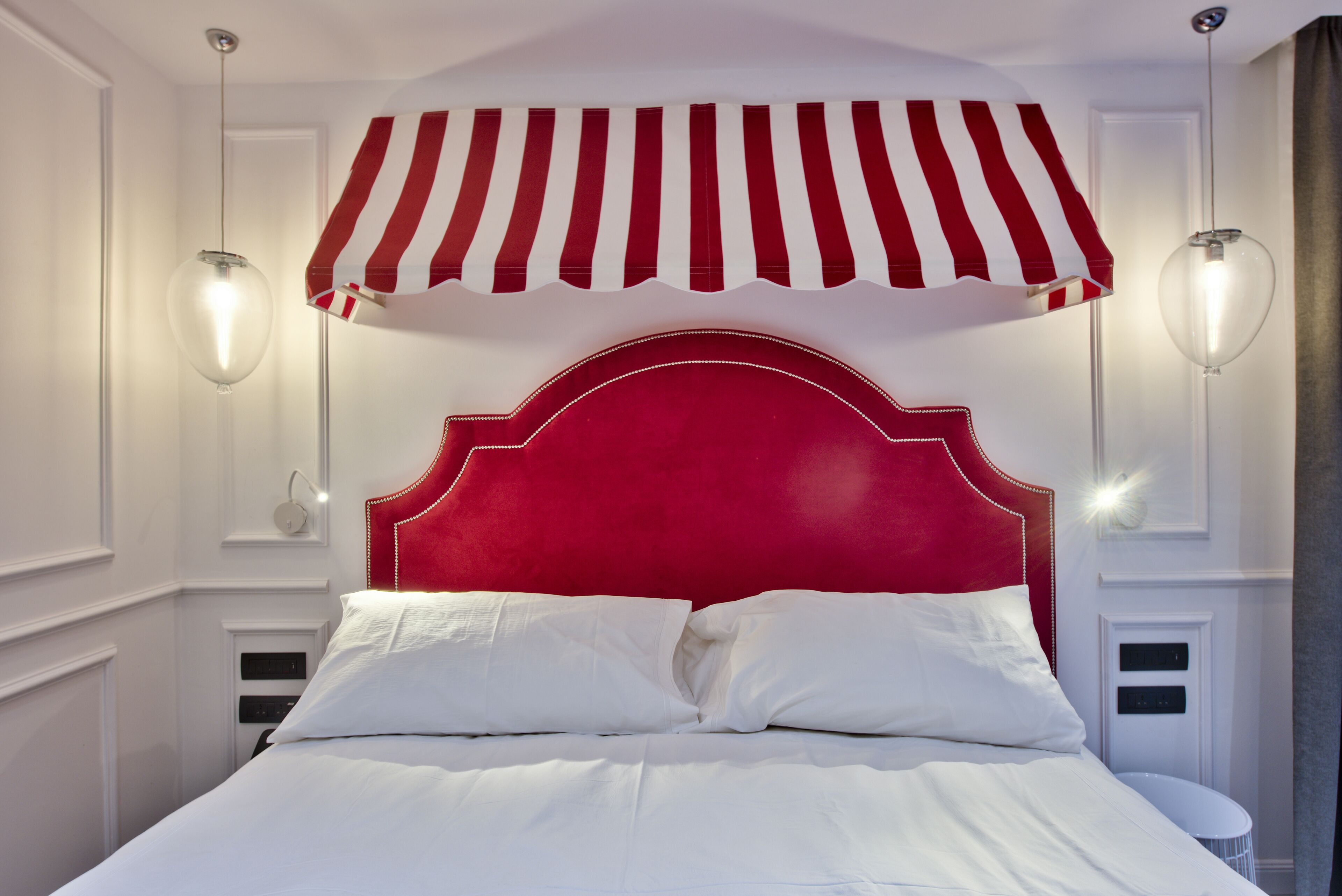 Una cama con cabecera roja, ropa de cama blanca y dos lámparas fijadas a la pared.