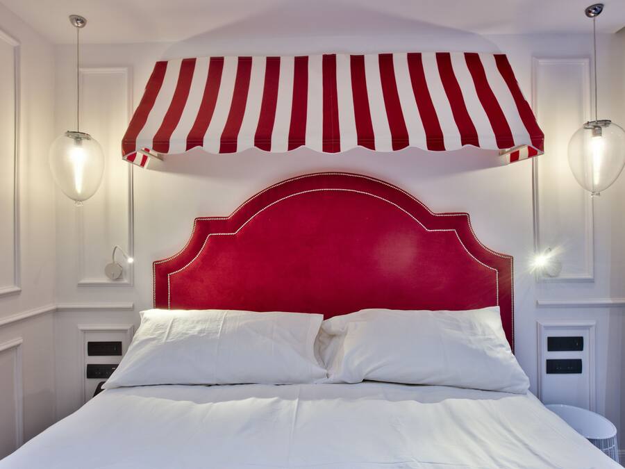Una cama con cabecera roja, ropa de cama blanca y dos lámparas fijadas a la pared.