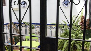 Blick durch ein Metallgitter auf einen Innenhof mit einem grünen Tisch und Stühlen, einer Wand mit blau-weißen Fliesenmustern und einem Blumentopf.