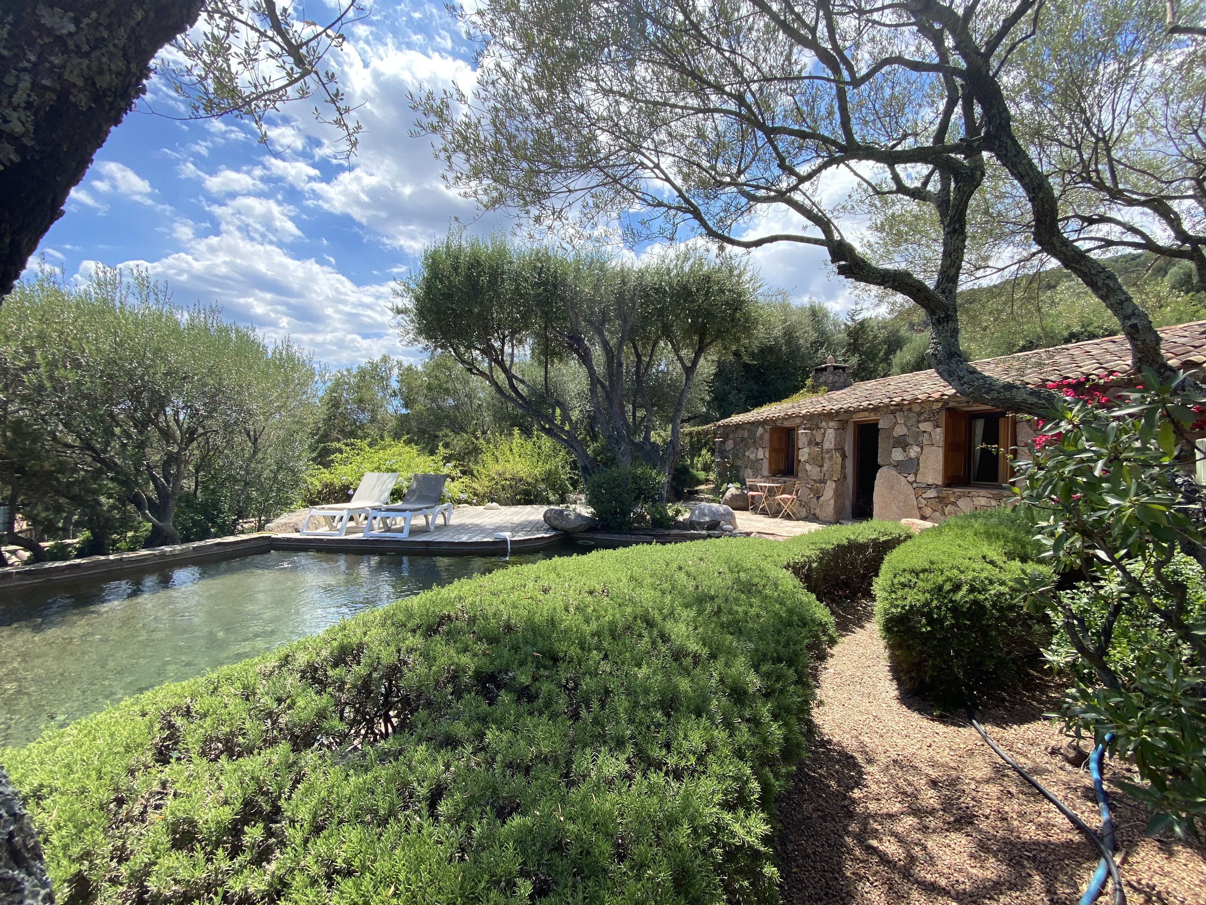 Une maison en pierre avec un toit en tuiles, une piscine et une terrasse en bois équipée de chaises longues.