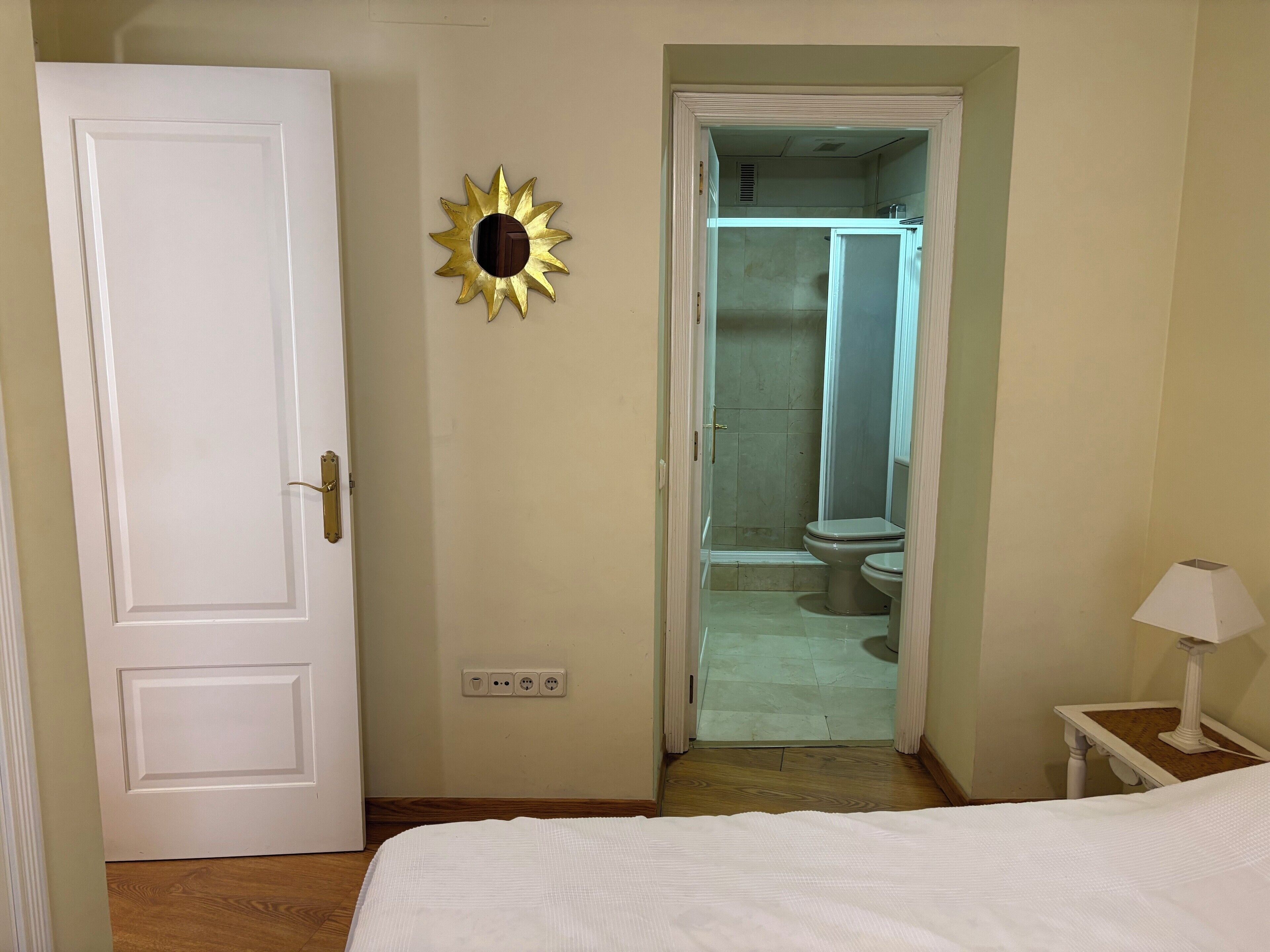 Una habitación con una cama, una puerta blanca, un espejo montado en la pared y un baño visible a través de una puerta abierta.