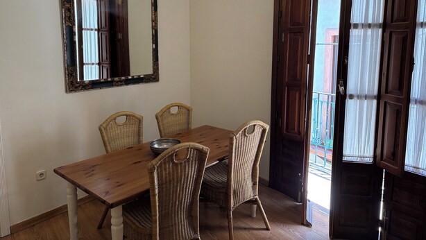 Un comedor con una mesa de madera y sillas de mimbre, un espejo grande en la pared y una puerta que da a un balcón exterior.