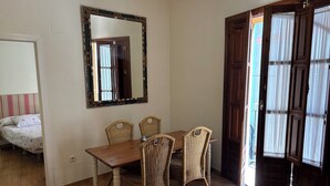 Un comedor con mesa de madera y sillas de mimbre, un espejo en la pared y una puerta entreabierta que da al exterior.