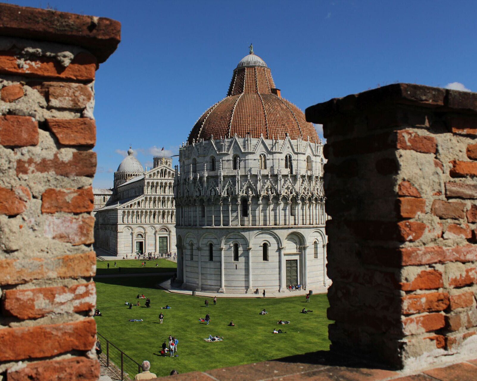A view of the Florence Cathedral's dome through a brick archway, with tourists on the surrounding lawn.