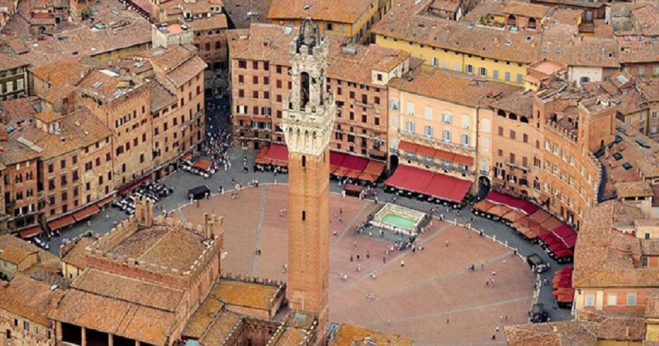 An aerial view of a historic square with a prominent tower, surrounded by traditional buildings with red-tiled roofs.