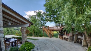 Un chemin de pierre mène à une terrasse en bois surmontée d’une pergola, le tout étant entouré d’une végétation luxuriante et agrémenté d’une cheminée en pierre.