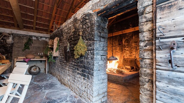 Une cheminée en pierre rustique avec un feu de bois, des poutres apparentes et un sol en pierre.