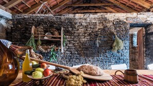 Une pièce rustique avec un mur en pierre, des poutres en bois au plafond et une table garnie de nourriture, ainsi qu’une cruche en bois.