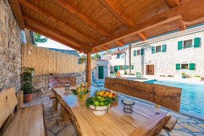 A wooden outdoor dining area with a stone wall, a pool, and a stone building with green shutters in the background.