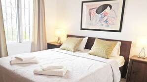 Una cama bien hecha con sábanas blancas y dos almohadas, una mesita de noche con una lámpara y una obra de arte enmarcada en la pared.