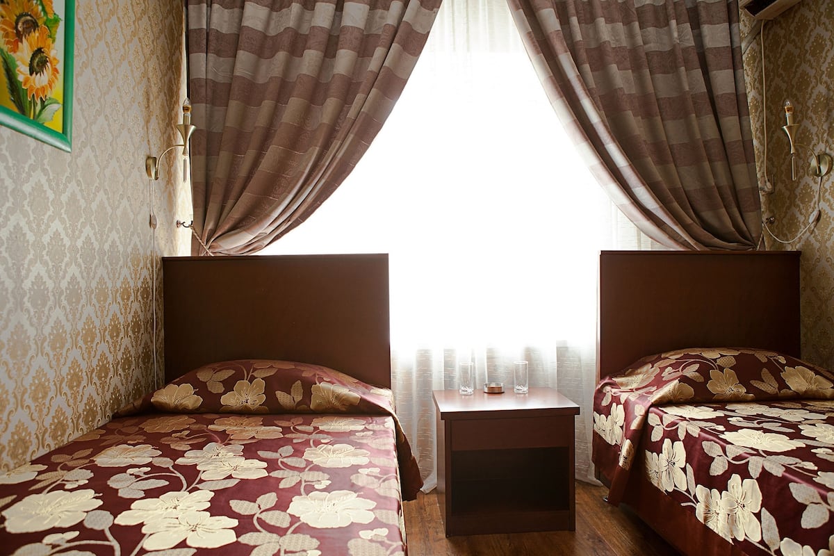 A room with two beds, each with a floral bedspread, a small bedside table with a candle, and a window with curtains.