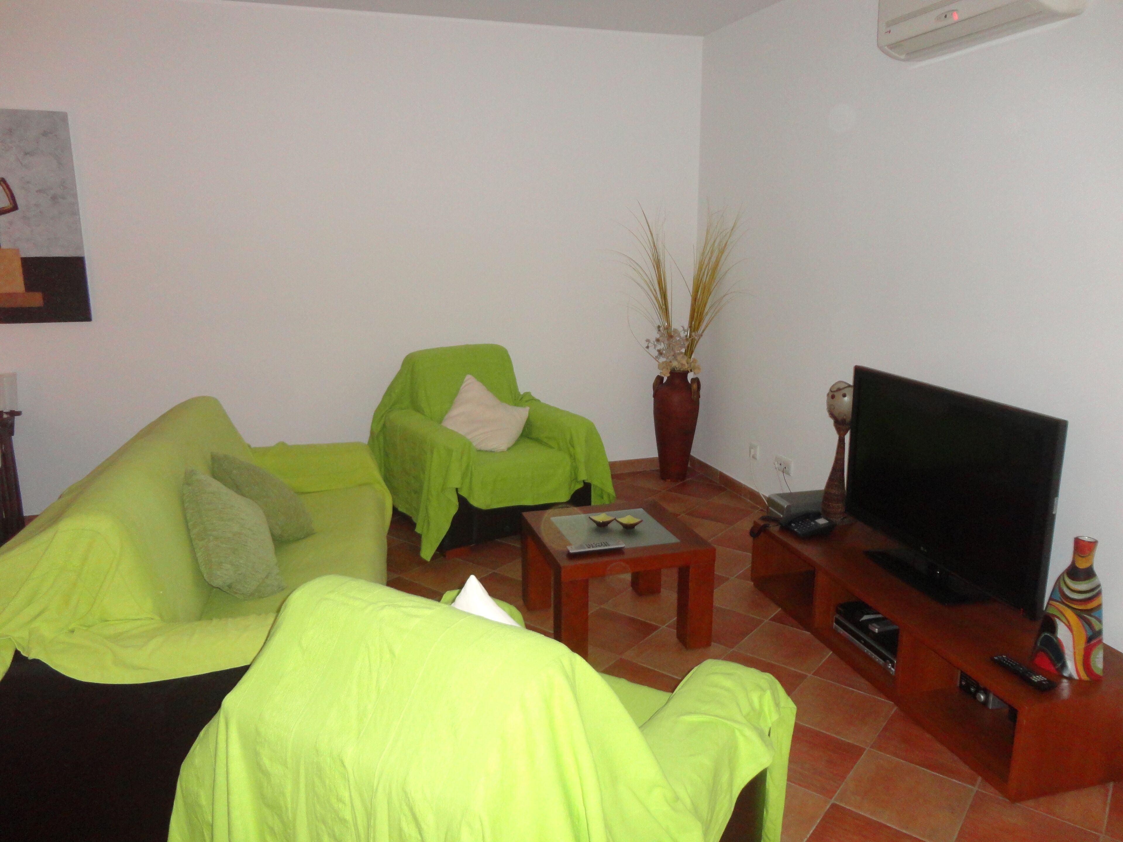 Una sala de estar con un sofá verde, un sillón verde más pequeño, una mesa de centro de madera, un mueble para televisión con un televisor de pantalla plana y una planta en maceta.