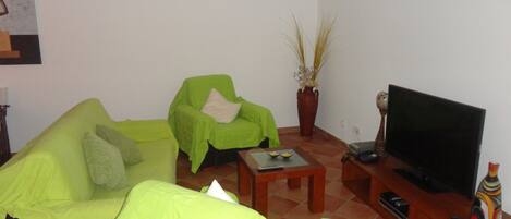 Una sala de estar con un sofá verde, un sillón verde más pequeño, una mesa de centro de madera, un mueble para televisión con un televisor de pantalla plana y una planta en maceta.