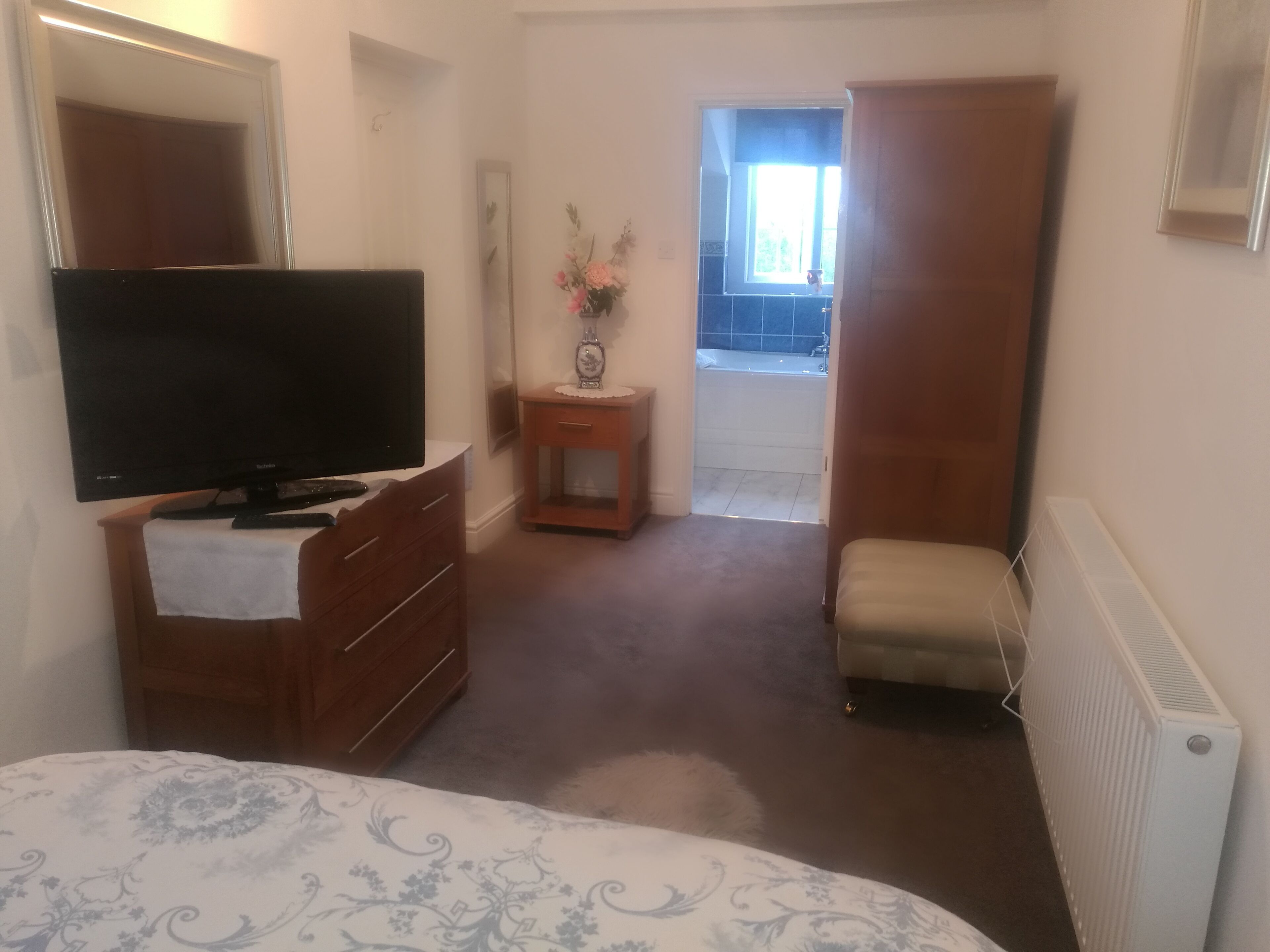 Et soveværelse med seng, fjernsyn, kommode, et lille bord med en vase, en radiator og en dør, der fører til en balkon.
