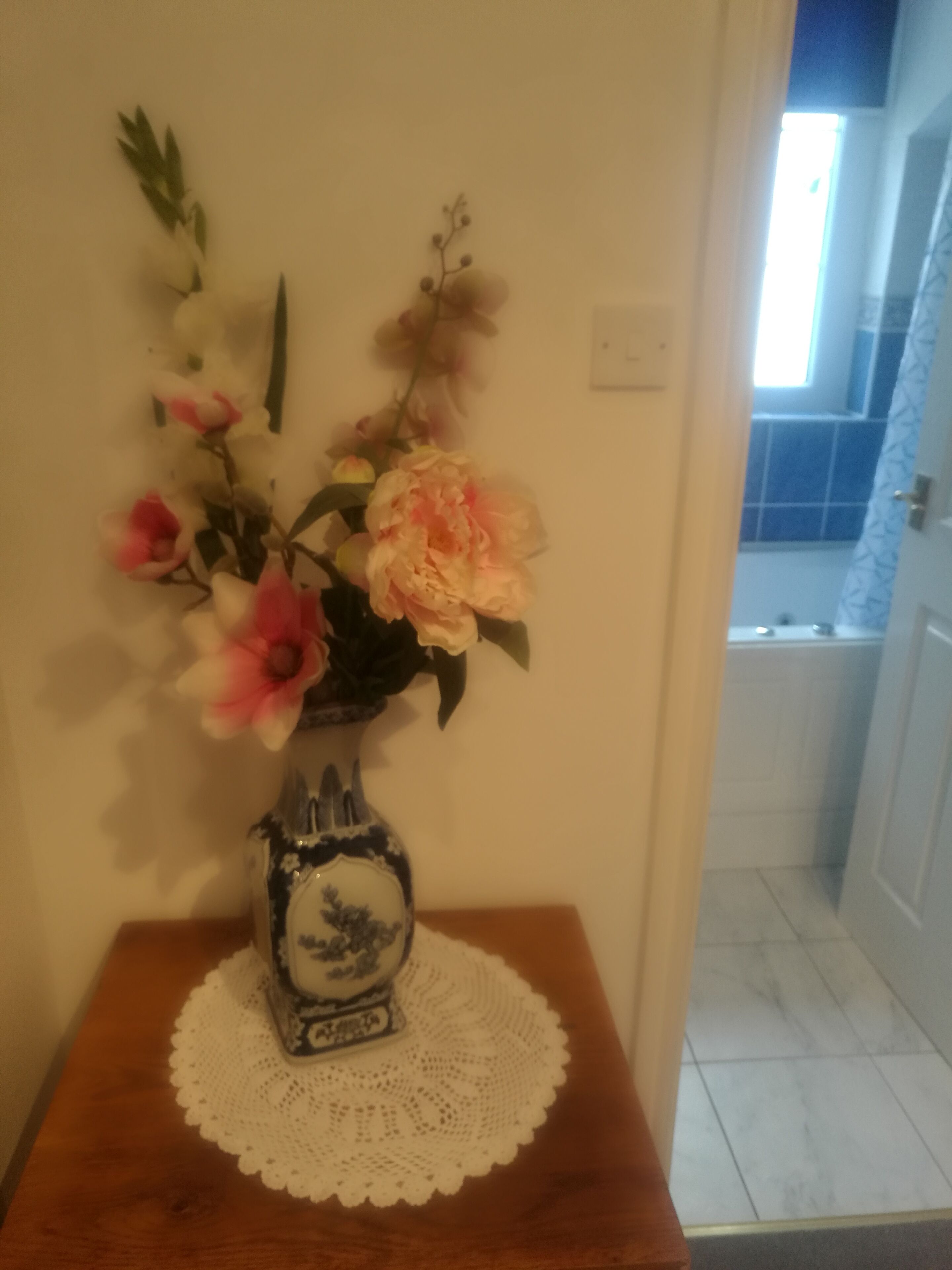 A vase with flowers on a wooden table with a doily, a white wall, and a door leading to a tiled area.