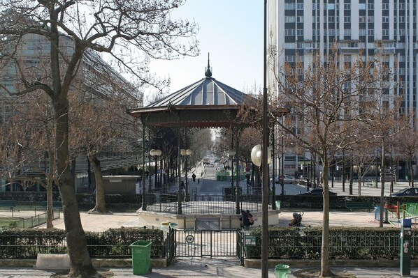 A gazebo in a park with a fountain in the background, surrounded by trees and a cityscape.