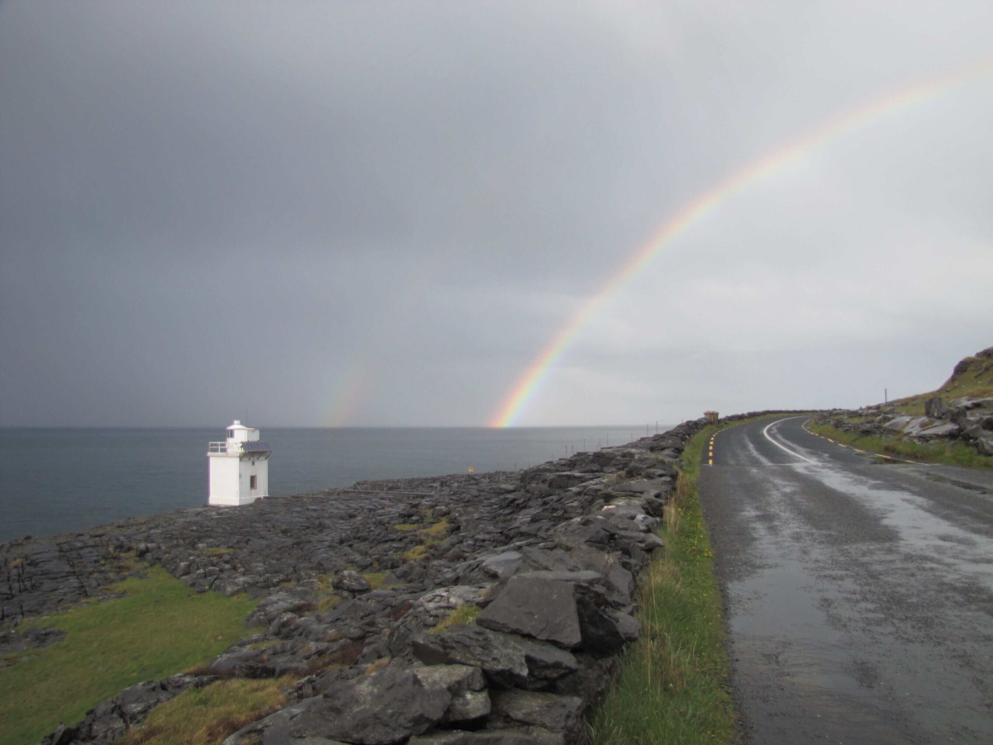 A coastal road with a lighthouse and a rainbow.