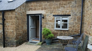 A stone building with a door, a window, a potted plant, and a small table with chairs. - Cosy Period Cottage In Unspoilt Village On The Edge Of The Cotswolds (North Newington)