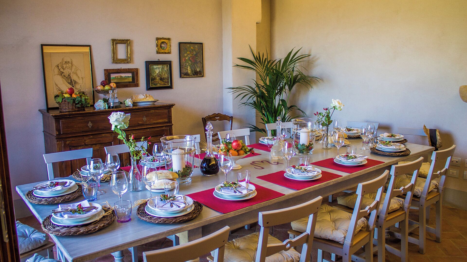 A dining room with a long table set for a meal, surrounded by chairs. There are framed pictures on the wall, a potted plant, and a wooden cabinet with decorative items.