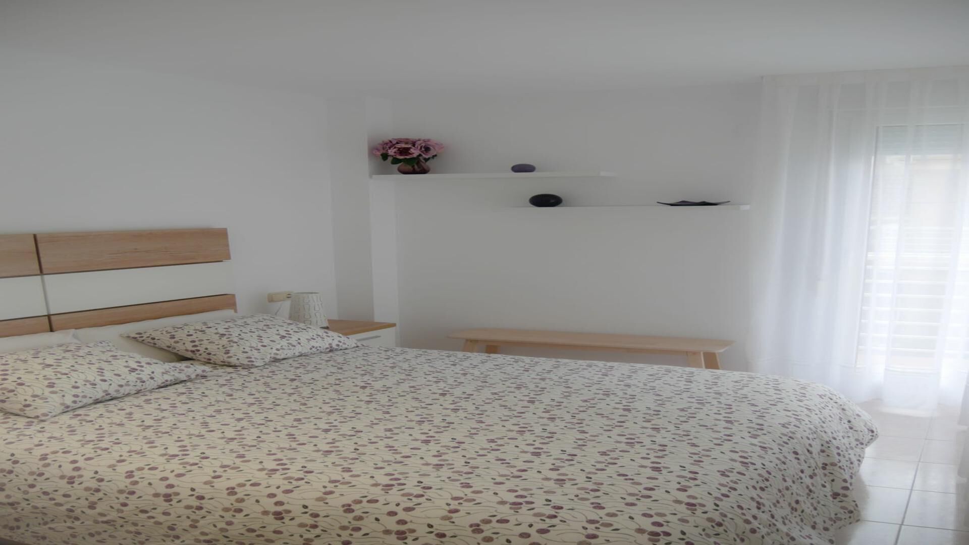 A bedroom with a bed, a bedside table, a nightstand, a bench, and a shelf with a vase and two decorative items.