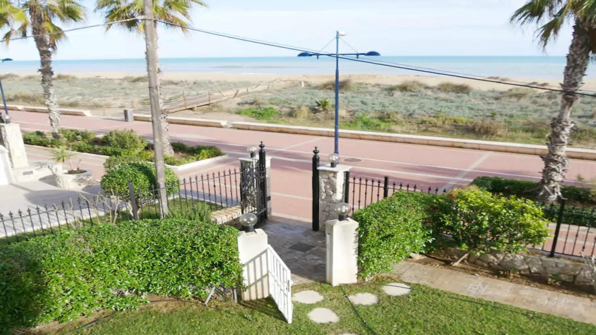 Een hekpoort met een pad dat uitkomt op een weg, omgeven door groen en palmbomen, met uitzicht op een strand.