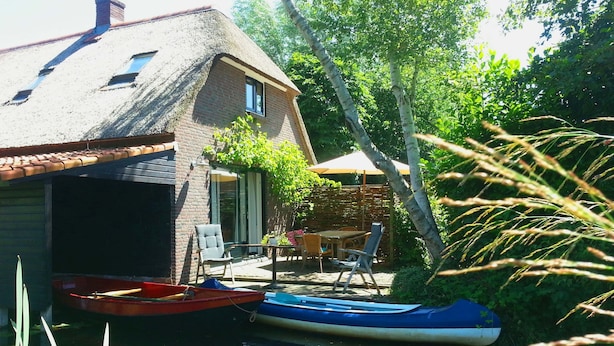 Une maison avec un toit de chaume, un petit quai avec deux bateaux et un patio aménagé avec du mobilier d’extérieur.