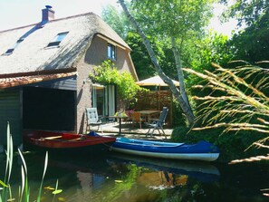 Een huis met rieten dak, een klein steiger met twee boten en een terras met tuinmeubilair.
