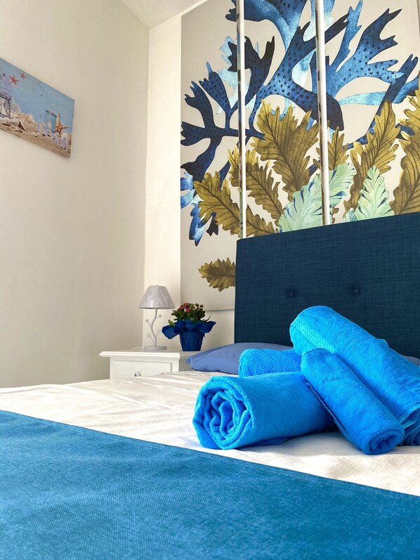 A neatly made bed with blue towels, a white nightstand with a lamp and a potted plant, and a wall with a leaf-patterned mural.