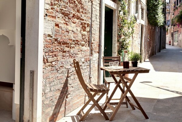 Un conjunto de mesa y sillas de madera al aire libre, frente a un muro de ladrillo, con una calle estrecha y edificios en el fondo.