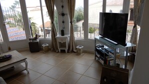 A living room with a TV, a sofa, a coffee table, and a small cabinet. There are large windows with curtains, and a balcony is visible outside. - Fantastic Villa Amare on top of the Montemar (Benissa)