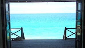 A room with a glass table and three cups, a view of the ocean through open doors, and a wooden deck with a railing.