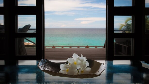 A room with a view of the ocean through a window. There is a bowl with white flowers on a table in the foreground.