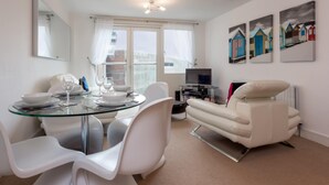 A modern living room with a glass dining table, white chairs, a white sofa, and a TV. There are three paintings on the wall and a window with curtains.