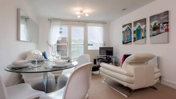 A modern living room with a glass dining table, white chairs, a white sofa, and a TV. There are three paintings on the wall and a window with curtains.