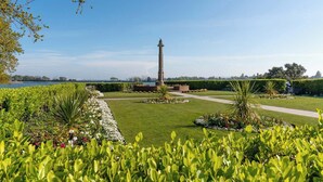 A well-maintained garden with a tall monument, neatly trimmed hedges, and a pathway leading to a body of water.