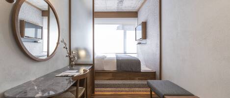 Un dormitorio moderno con una cama, un banco de madera, un escritorio con una silla, un espejo redondo y un televisor en la pared.