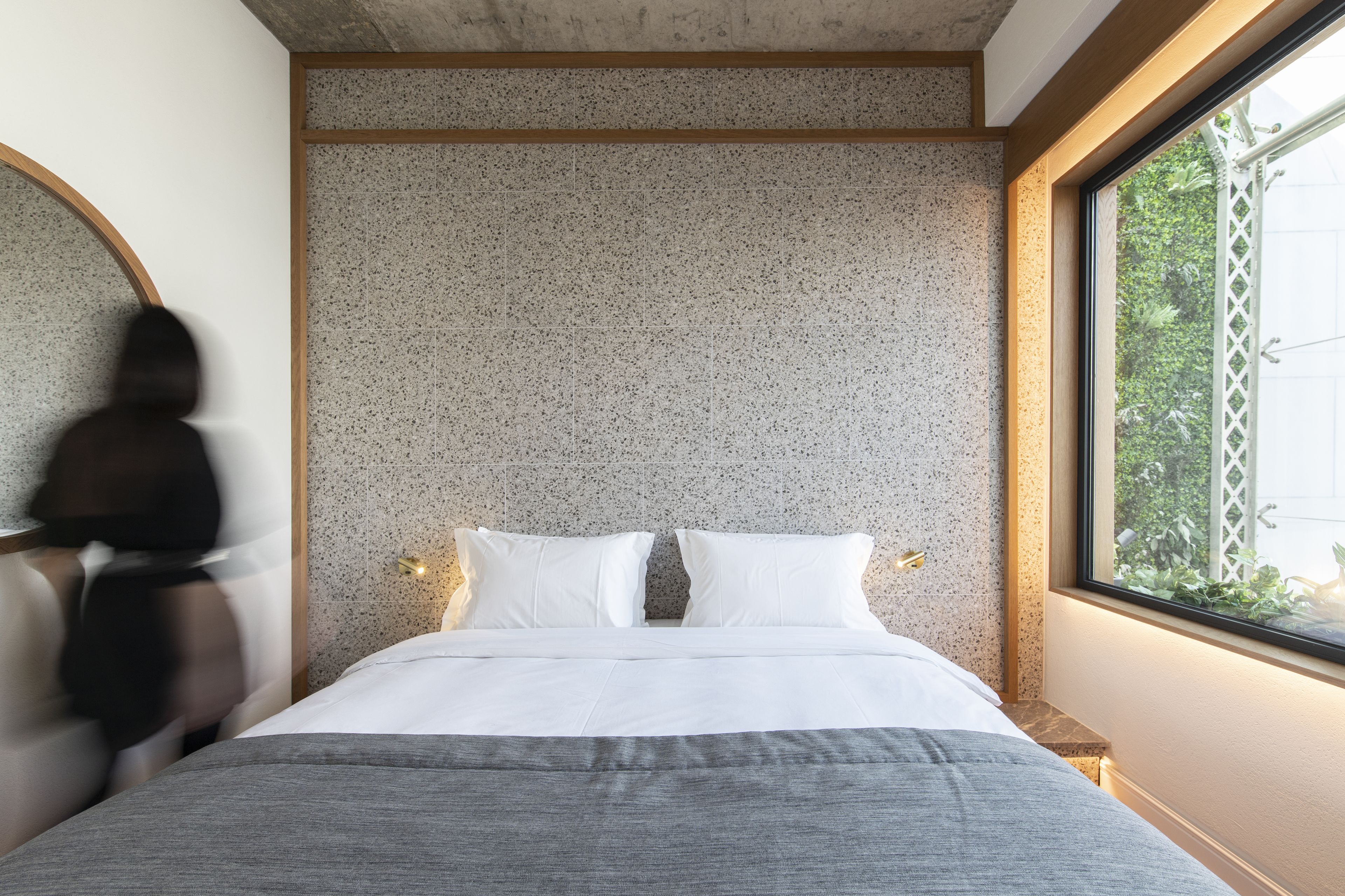 A modern bedroom with a large bed, a round mirror, and a window with a view of greenery.