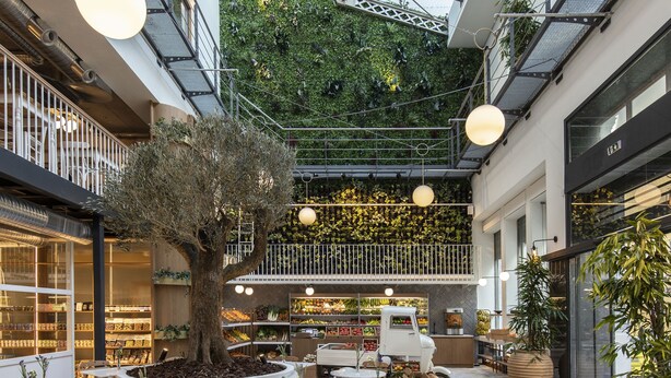 An indoor courtyard with tables and chairs, a central tree, and a food display. - Ergon House Athens (Athens)