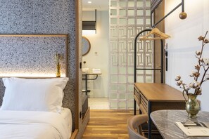 Quarto de hotel com cama, escrivaninha de madeira, um vaso com flores secas e um suporte de toalhas fixado na parede.
