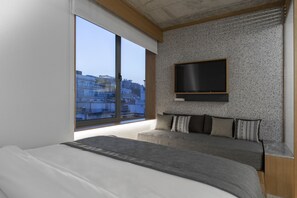 Quarto moderno com uma cama grande, televisão na parede e vista para edifícios por uma janela ampla.