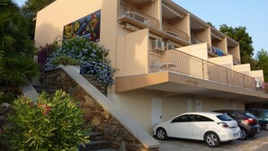 Un edificio a più piani con una scala in pietra che conduce a un balcone. Davanti, auto parcheggiate e una facciata beige con un murale su un lato.