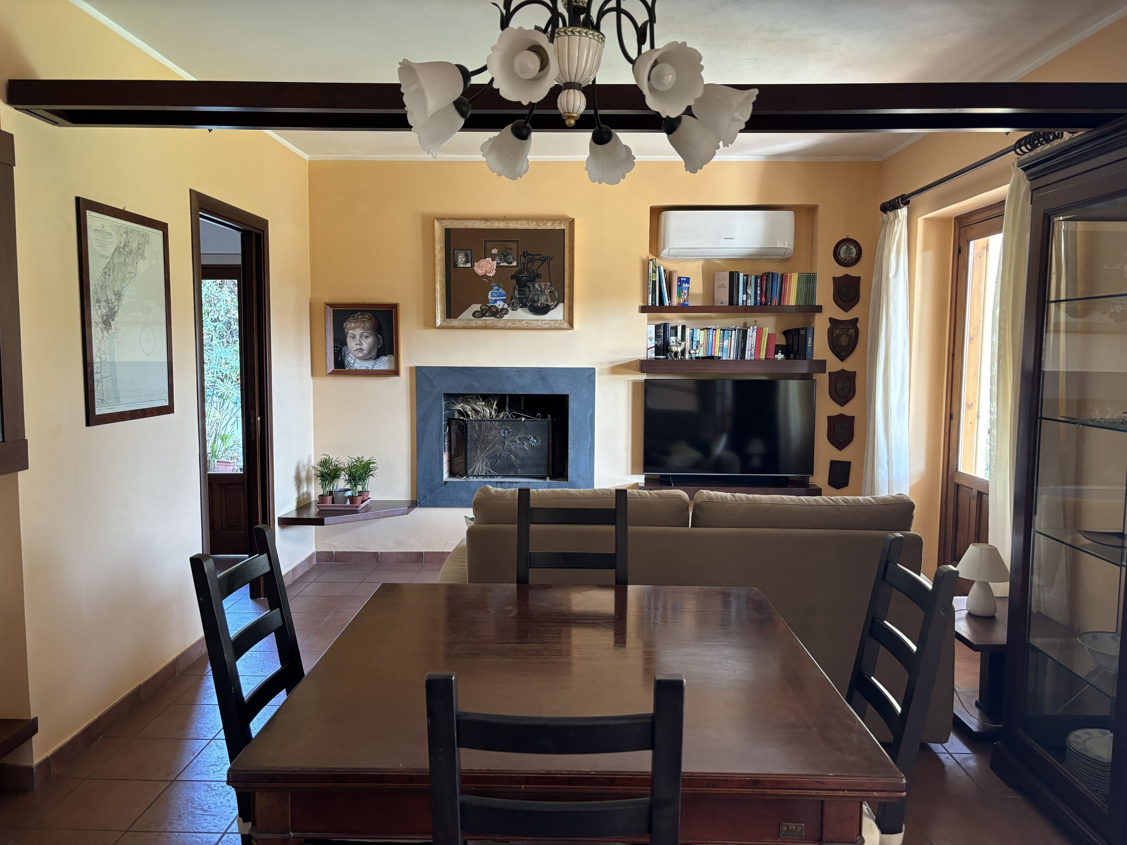 Un comedor con mesa y sillas de madera, chimenea, sofá y estantería con libros y objetos decorativos.