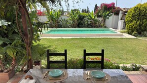 Ein Esstisch mit zwei Stühlen im Freien, im Hintergrund ein Pool und üppiges Grün.